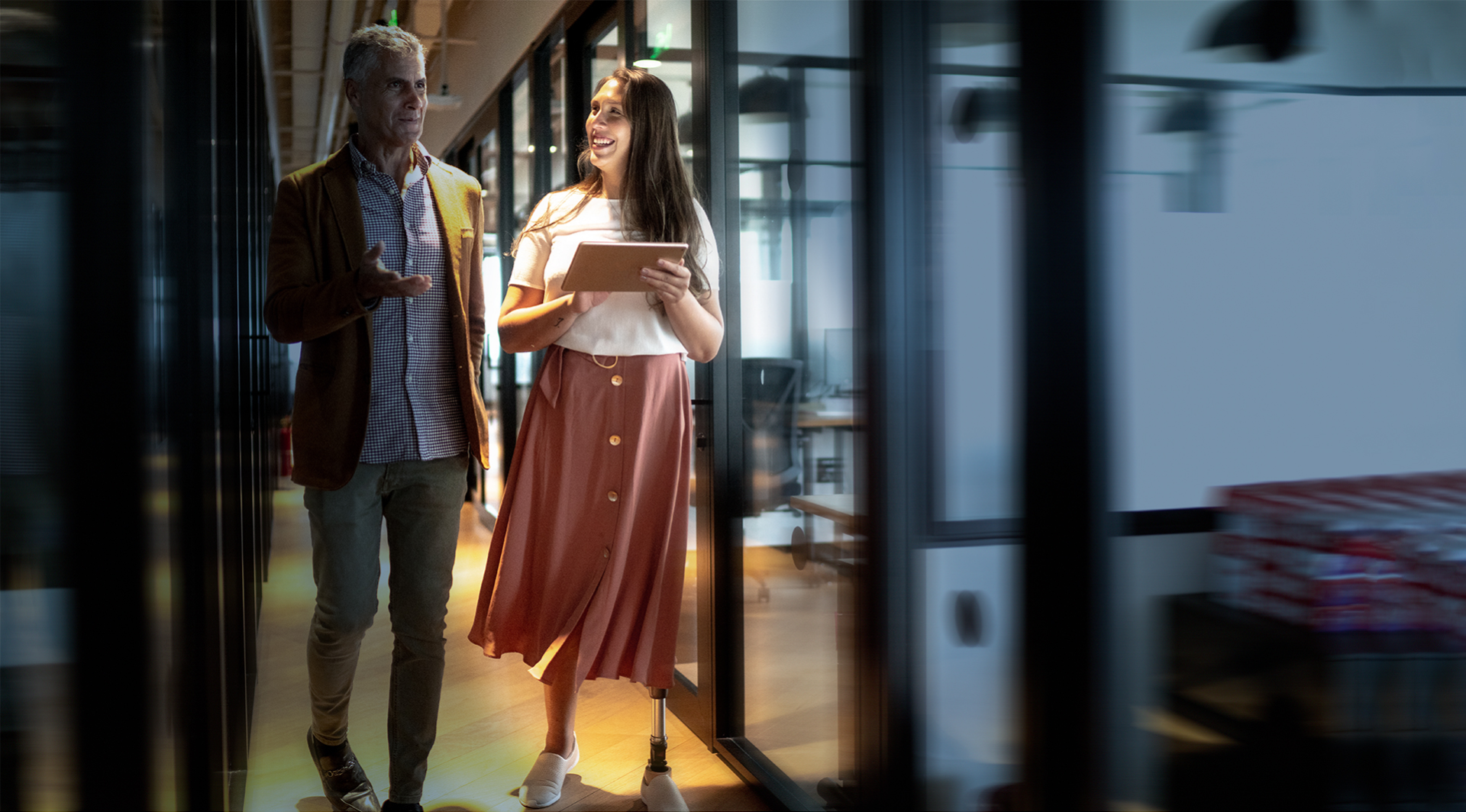 Ein Mann und eine Frau gehen in einem Gang zwischen verglasten Büros und unterhalten sich. Die Frau hat eine Beinprothese und hält ein Tablet.