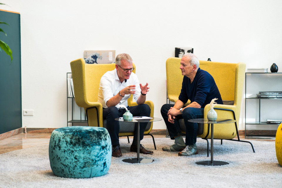 Hr. Hagen trägt ein weißes Hemd mit schwarzer Hose. Hr. Otto-Albrecht trägt dunkelblaue Kleidung. Beide sitzen in einem hellen Raum in gelben Sesseln und unterhalten sich.