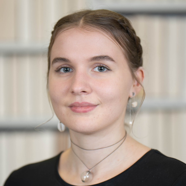 Linda Diekmann trägt ein schwarzes Oberteil, ihr Haar ist zum Zopf gebunden. Sie trägt Ohrringe und eine enge Halskette.