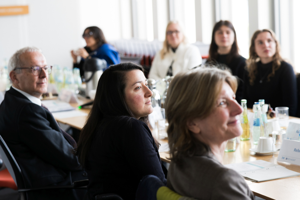 6 Frauen und 1 Mann sitzen an einem Tisch mit Blick nach vorne und hören einem Vortrag zu.