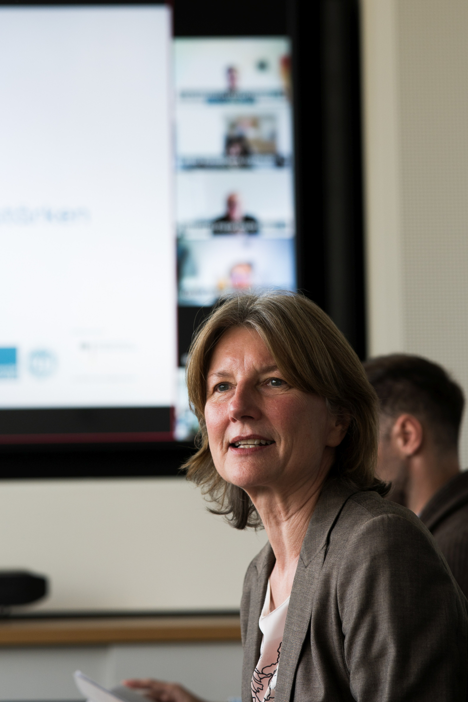 Prof. Dr. Mathilde Niehaus blickt nach links und referiert. Im Hintergrund sind eine Leinwand und eine Person verschwommen zu sehen.