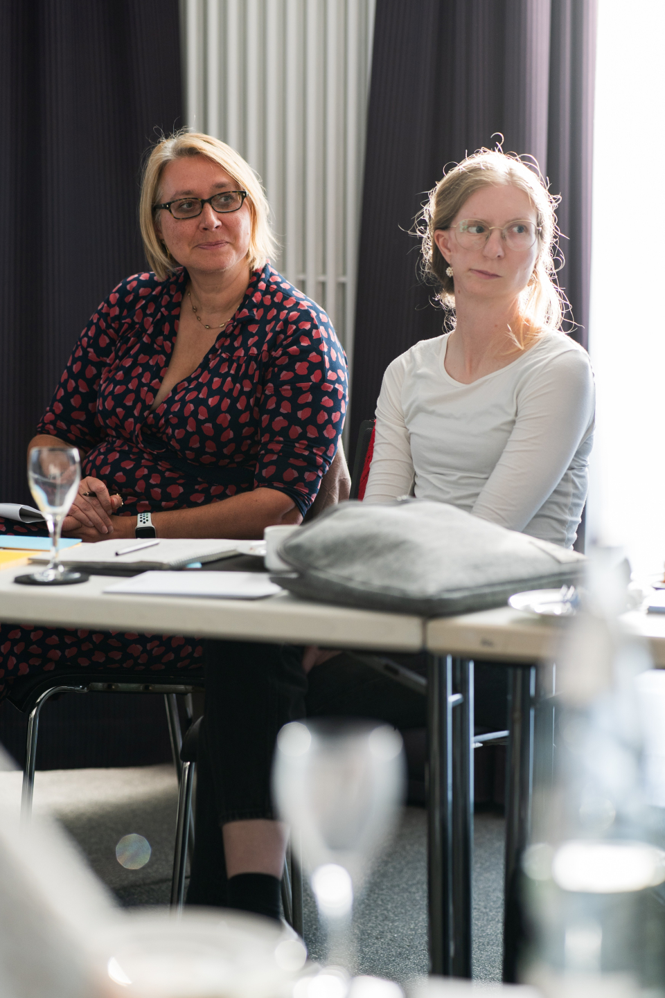 Fr. Bellen (links) und Fr. Rhein (rechts) sitzen an einem Tisch und blicken nach links. Sie verfolgen eine Präsentation.