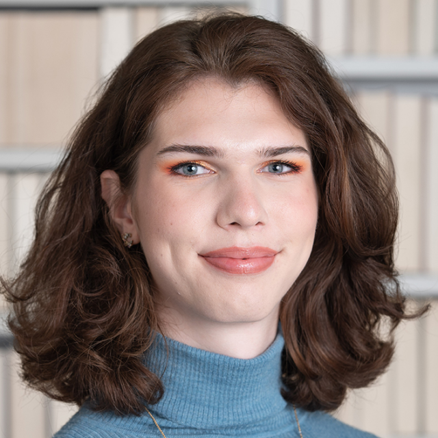 Aeyla Katzenbach hat braunes, schulterlanges Haar. Sie trägt einen blauen Rollkragenpullover und lächelt in die Kamera. Der Hintergrund ist unscharf.