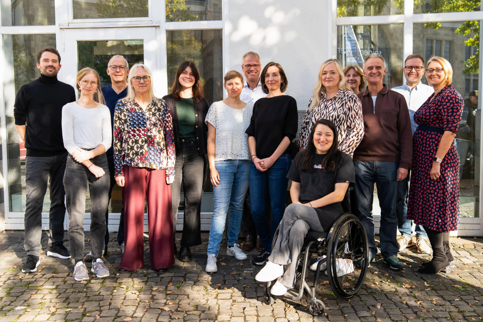 5 Männer und 9 Frauen (eine davon im Rollstuhl) stehen vor einem Gebäude für ein Gruppenbild. 