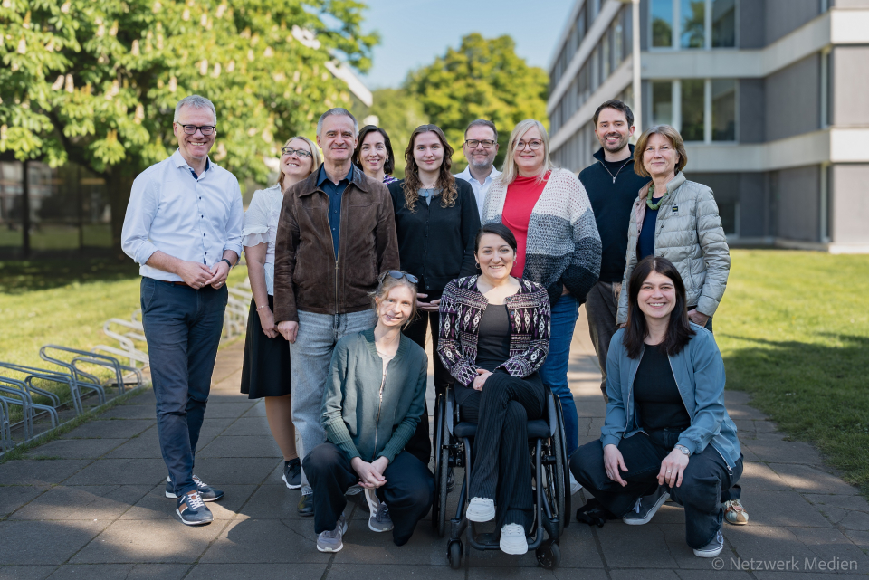 Das Projektteam, 8 Frauen und 4 Männer, auf dem Campus der Universität zu Köln zwischen Bäumen und einem Gebäude. Eine Person sitzt im Rollstuhl, die anderen stehen und knien.