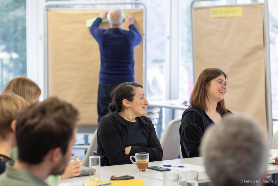 6 Personen sitzen an Tischen. Zwei Frauen lachen nach links. Im Hintergrund hängt ein Mann Notizen an einer Pinnwand auf. 6 Personen sitzen an Tischen. Zwei Frauen lachen nach links. Im Hintergrund hängt ein Mann Notizen an einer Pinnwand auf.